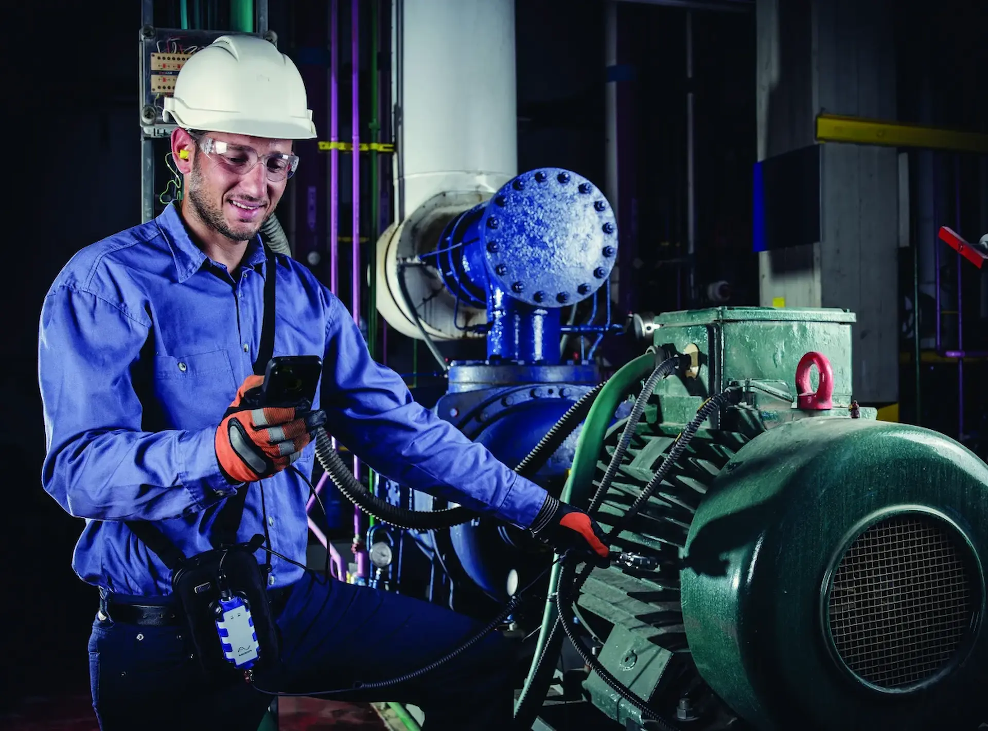 A worker in safety gear checks his phone while standing beside industrial machinery