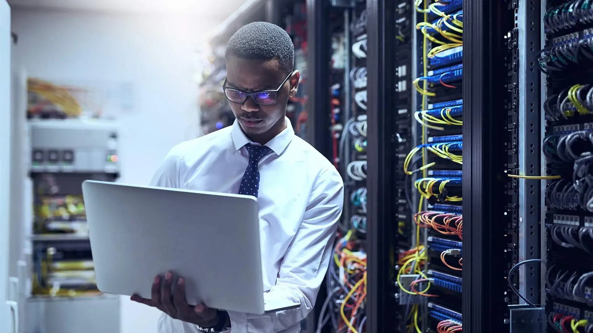 A man in formal attire analyzes data on a laptop amidst server racks