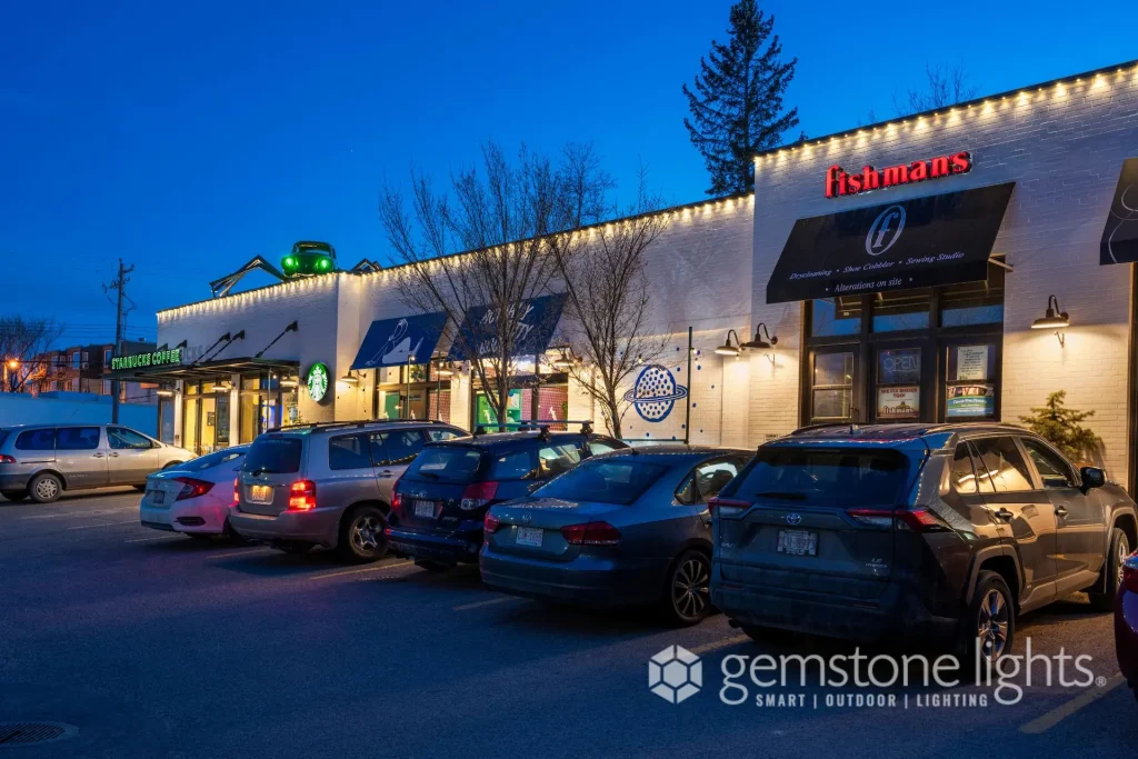 A row of brightly lit shops lines a street at twilight, with parked cars nearby