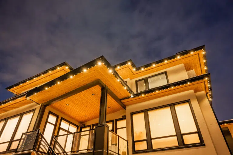 A modern house shines under a dusky sky, adorned with warm string lights